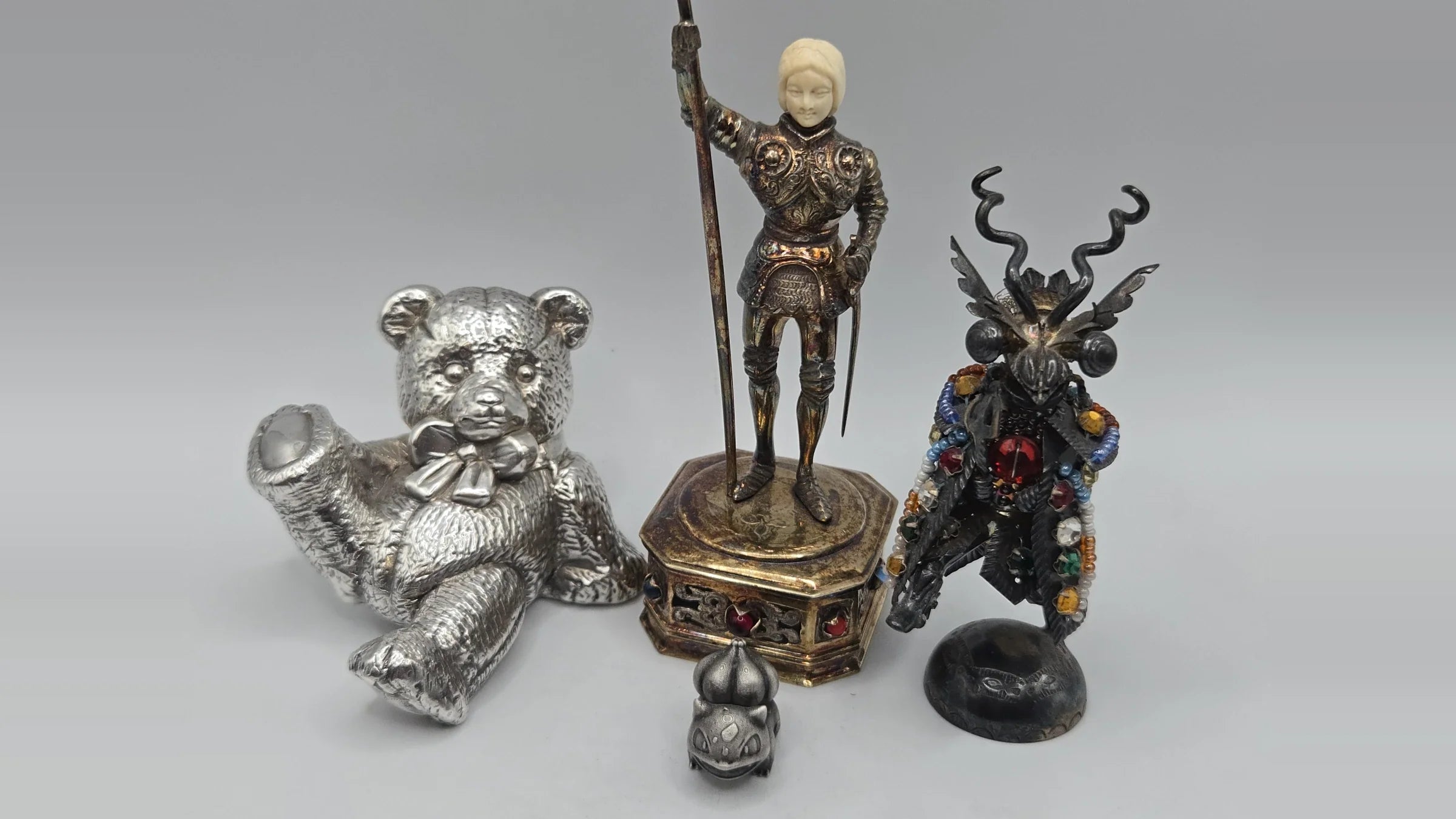 Five silver figurines from the Better Than Bullion collection showing different surface states — a mirror-bright teddy bear, an iridescently toned armoured knight on a gilt base, a black-patinated jewelled figure, and a matte grey character figurine